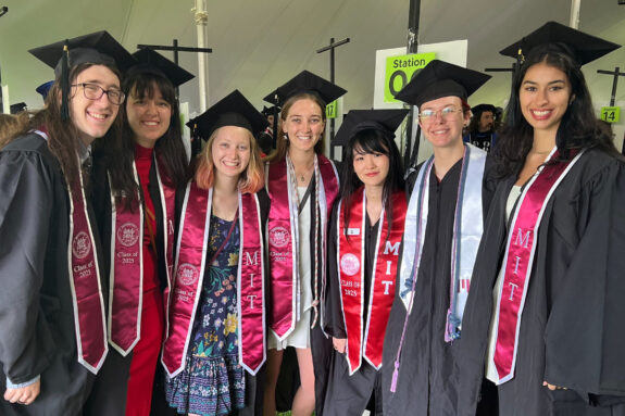 Seven undergraduates gathered wearing commencement caps and gowns under a tent.