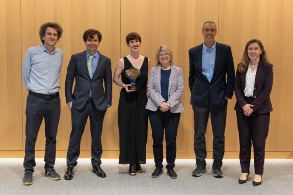 Nikos Trichakis, Caspar Hare, Annaliese Meyer, Sally Kornbluth, Dan Huttenlocher, and Asu Ozdaglar stand in a row in front of a light wooden wall. Meyer holds a colorful glass award.